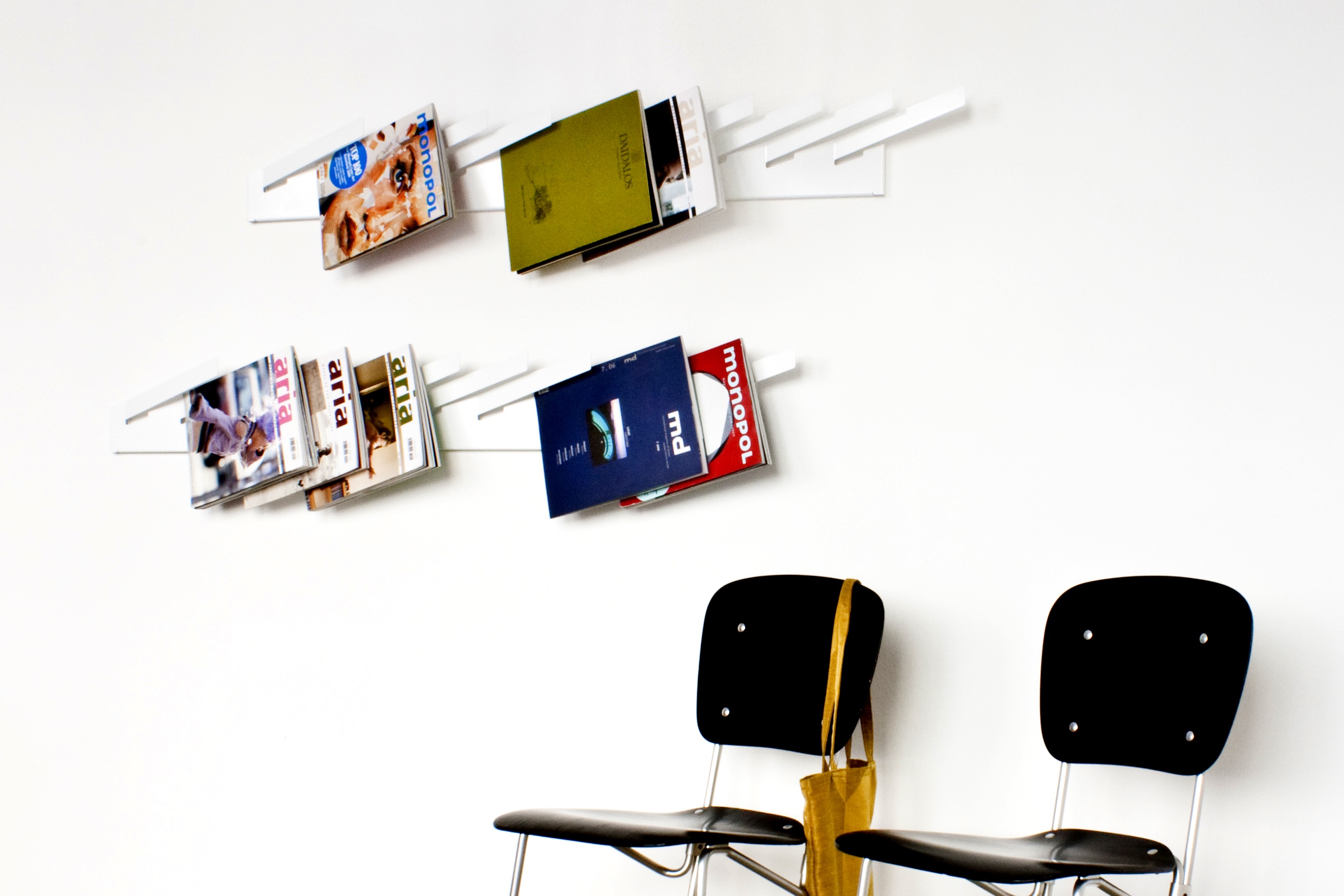 Weißes Wandregal mit Zeitschriften und zwei schwarzen Stühlen mit gelber Tasche vor weißer Wand.