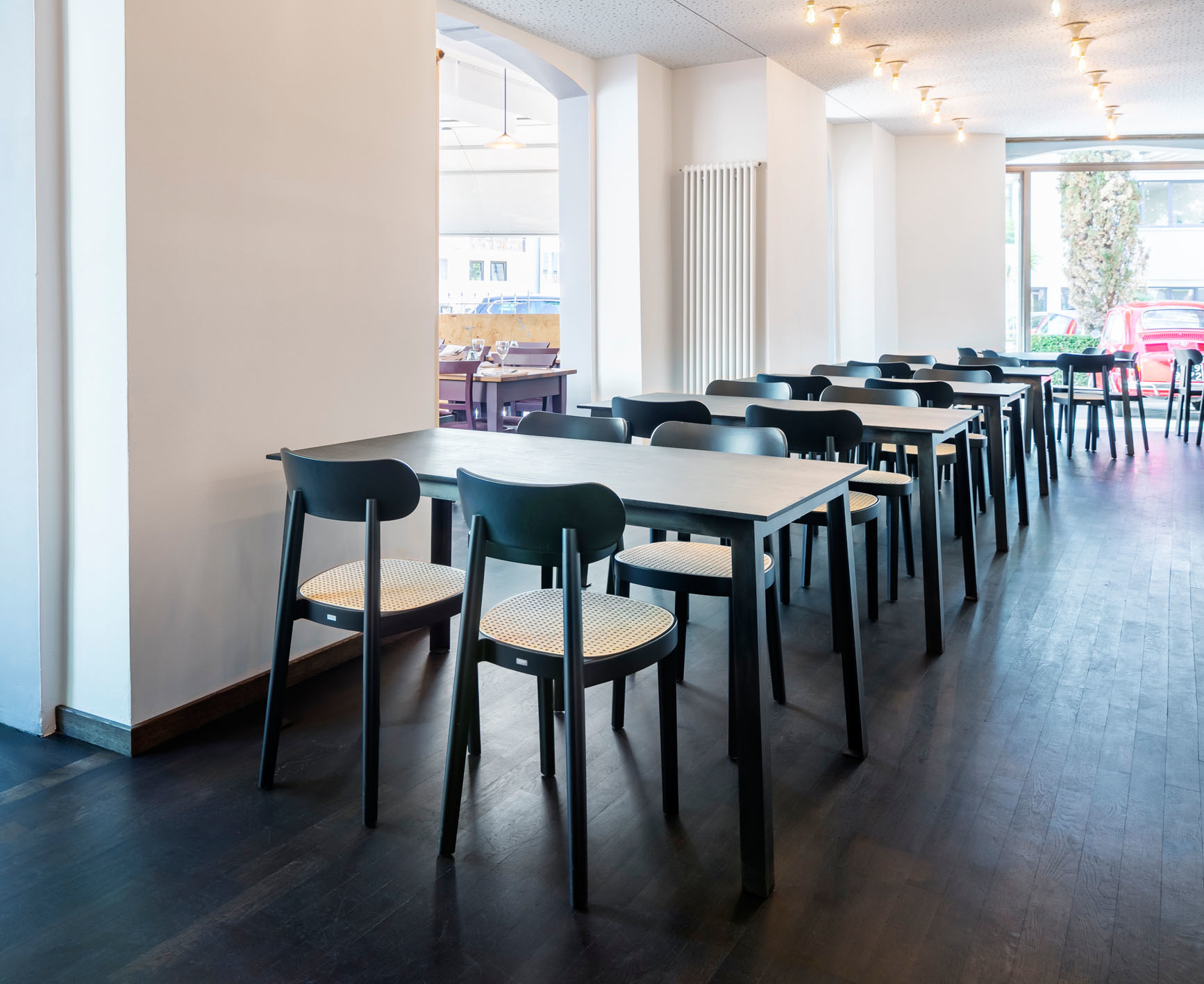 Reihe von schwarzen Holzstühlen vom Typ Thonet 118 mit Rohrgeflecht-Sitzfläche an Tischen in Restaurant.