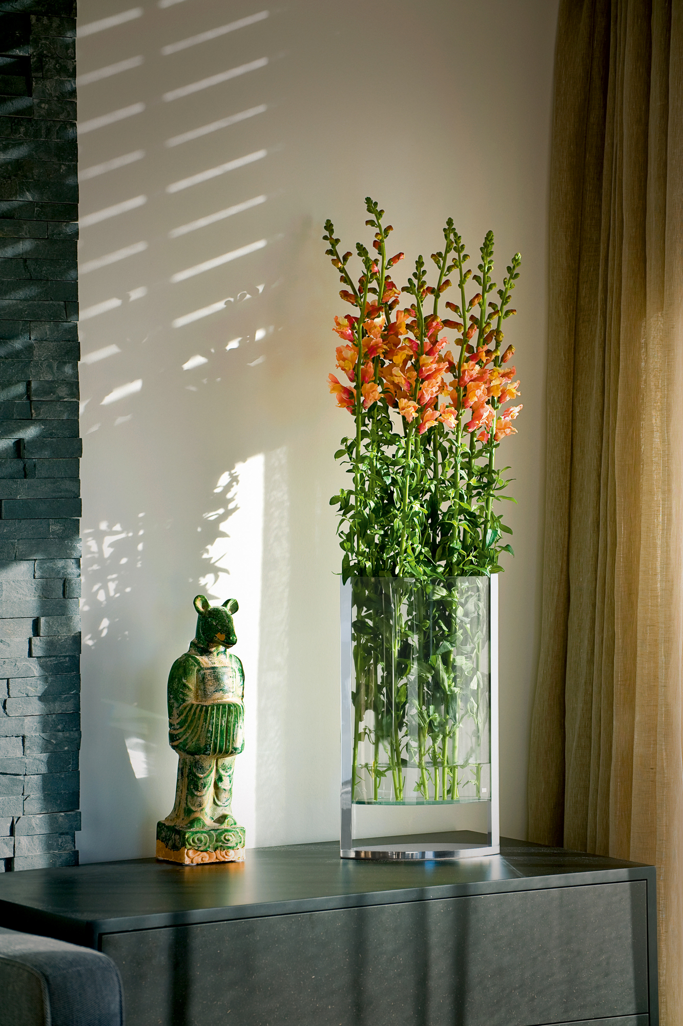 Decade Vase S Philippi mit orangefarbenen Blumen auf einer Kommode, neben einer grünen Statue.