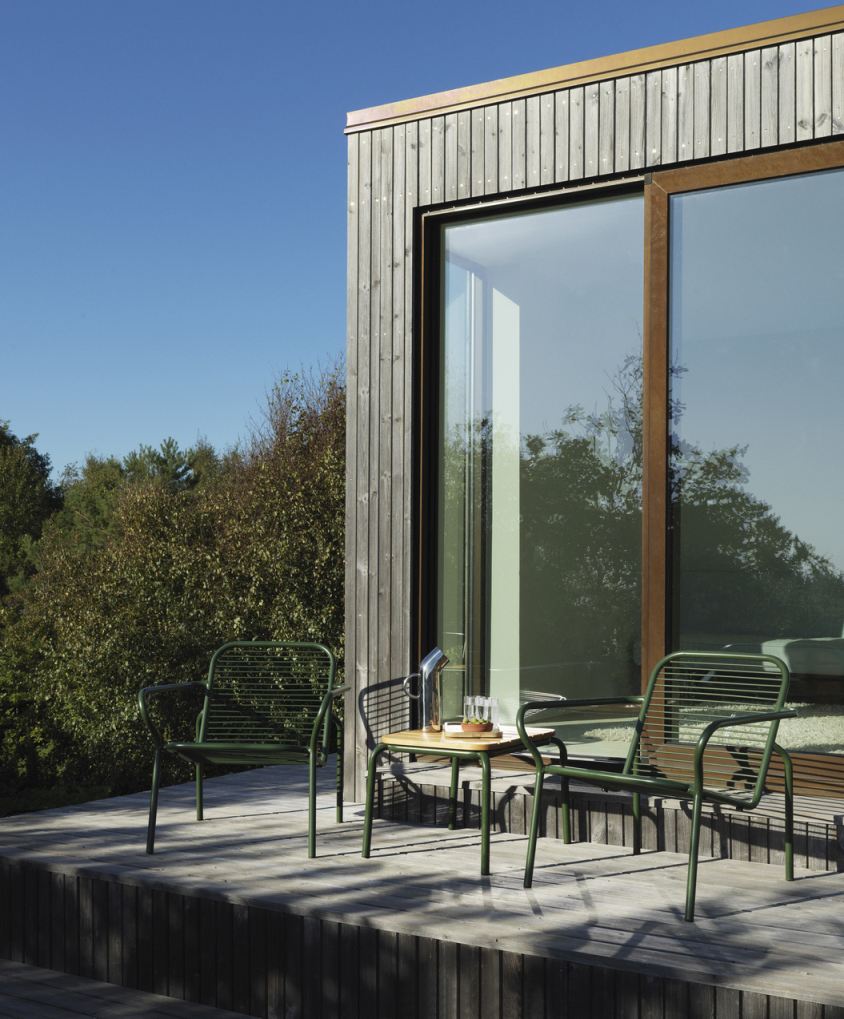 Normann Copenhagen Vig Table Outdoor mit zwei grünen Stühlen auf Holzterrasse vor modernem Haus.