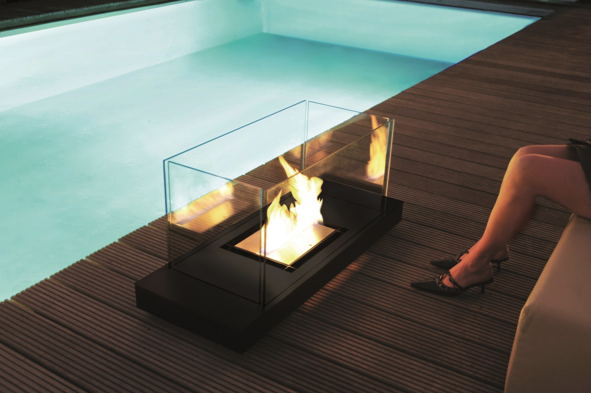 Uni Flame Ethanol Kamin Radius neben Pool auf Holzdeck mit Frau auf Liege.
