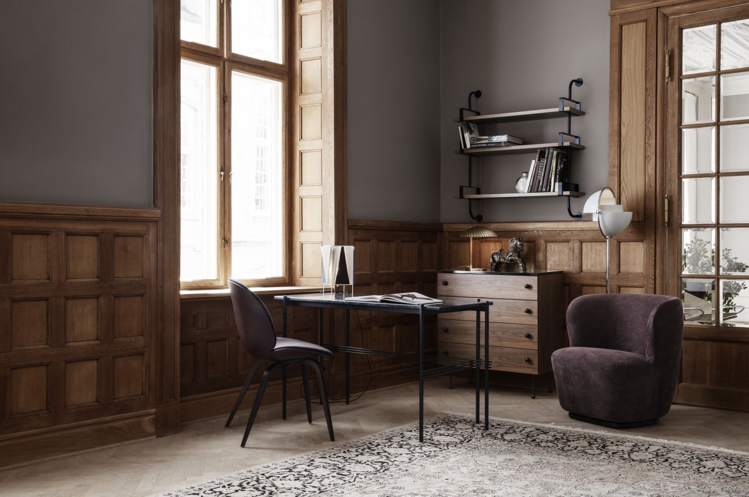 Elegantes Interieur mit Gubi Kommode, Schreibtisch, Sessel und Bücherregal vor einer holzvertäfelten Wand.