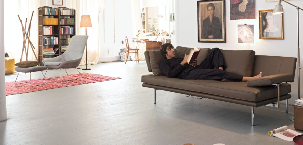 Mann liest auf braunem Walter Knoll LIVING PLATFORM Sofa mit Funktionsarmlehnen in modernem Wohnzimmer.