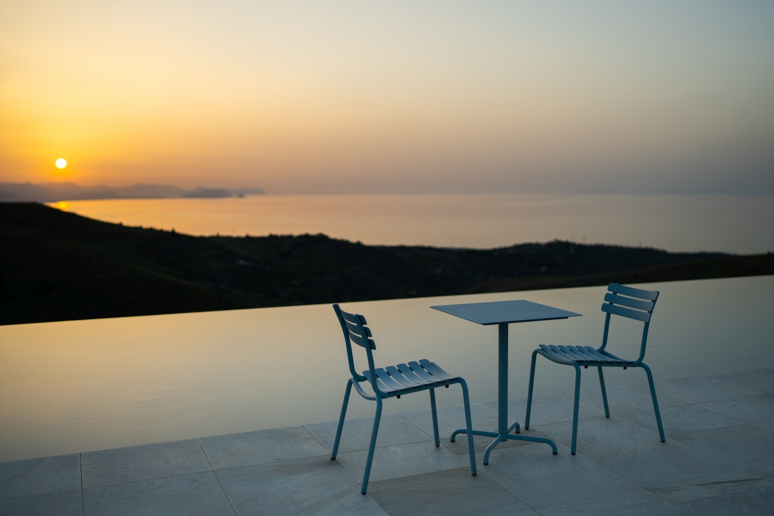 Alua Dining Chair von Houe: Blauer Outdoor-Stuhl und Tisch mit Meerblick bei Sonnenuntergang.