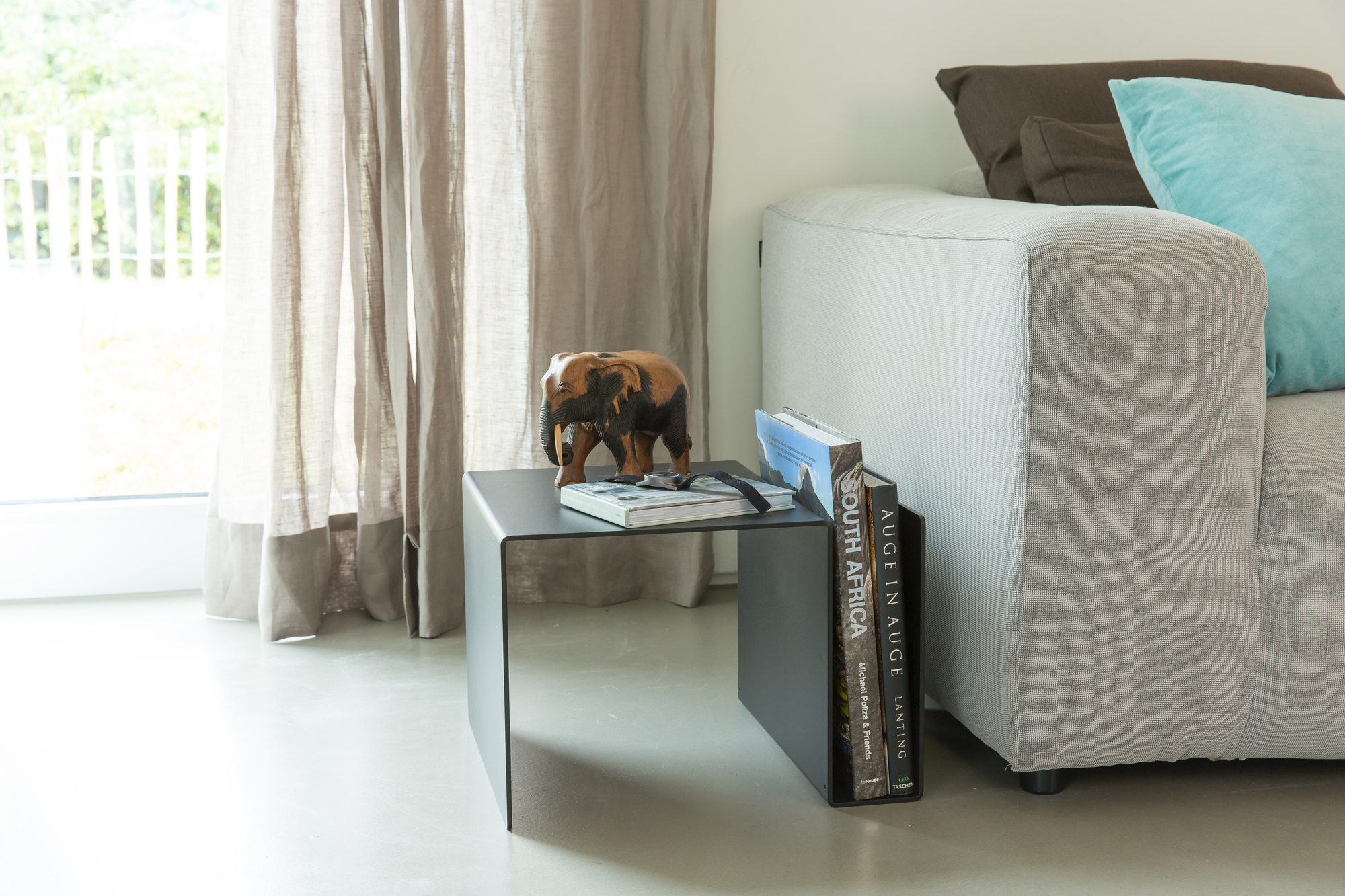 Huk Beistelltisch von Müller Möbelwerkstätten in Anthrazit mit Dekoration und Büchern neben einem Sofa.