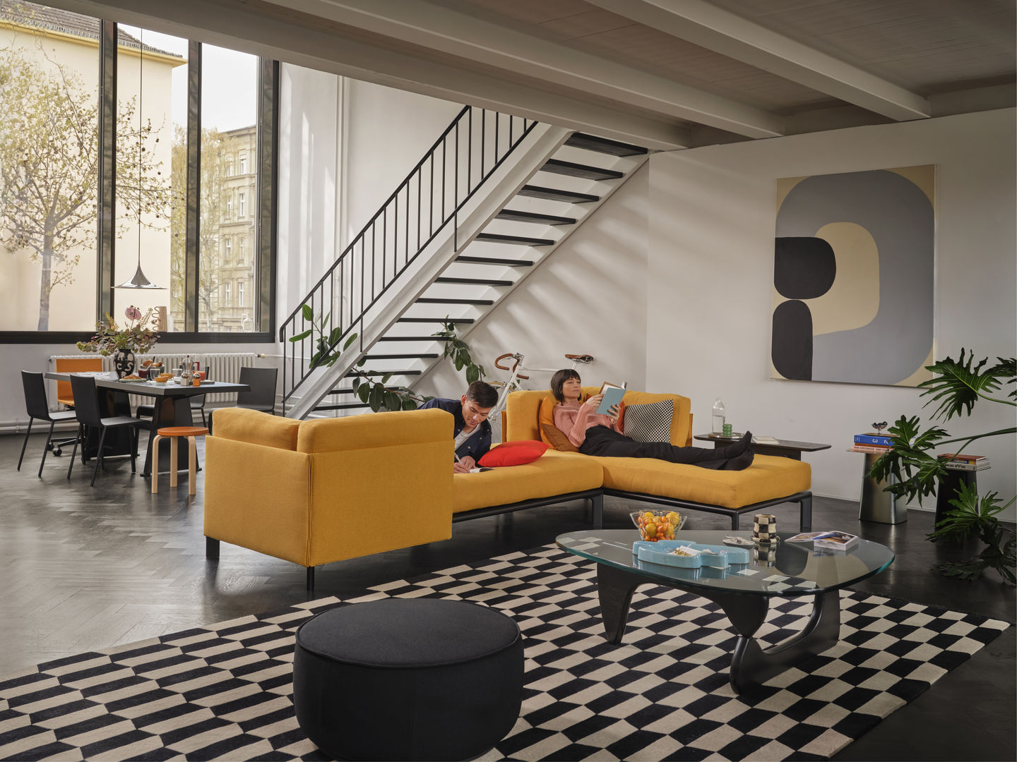 Wohnzimmer mit gelbem Anagram Sofa von Vitra, Personen lesen, moderner Inneneinrichtung und Schachbrettmuster-Teppich.
