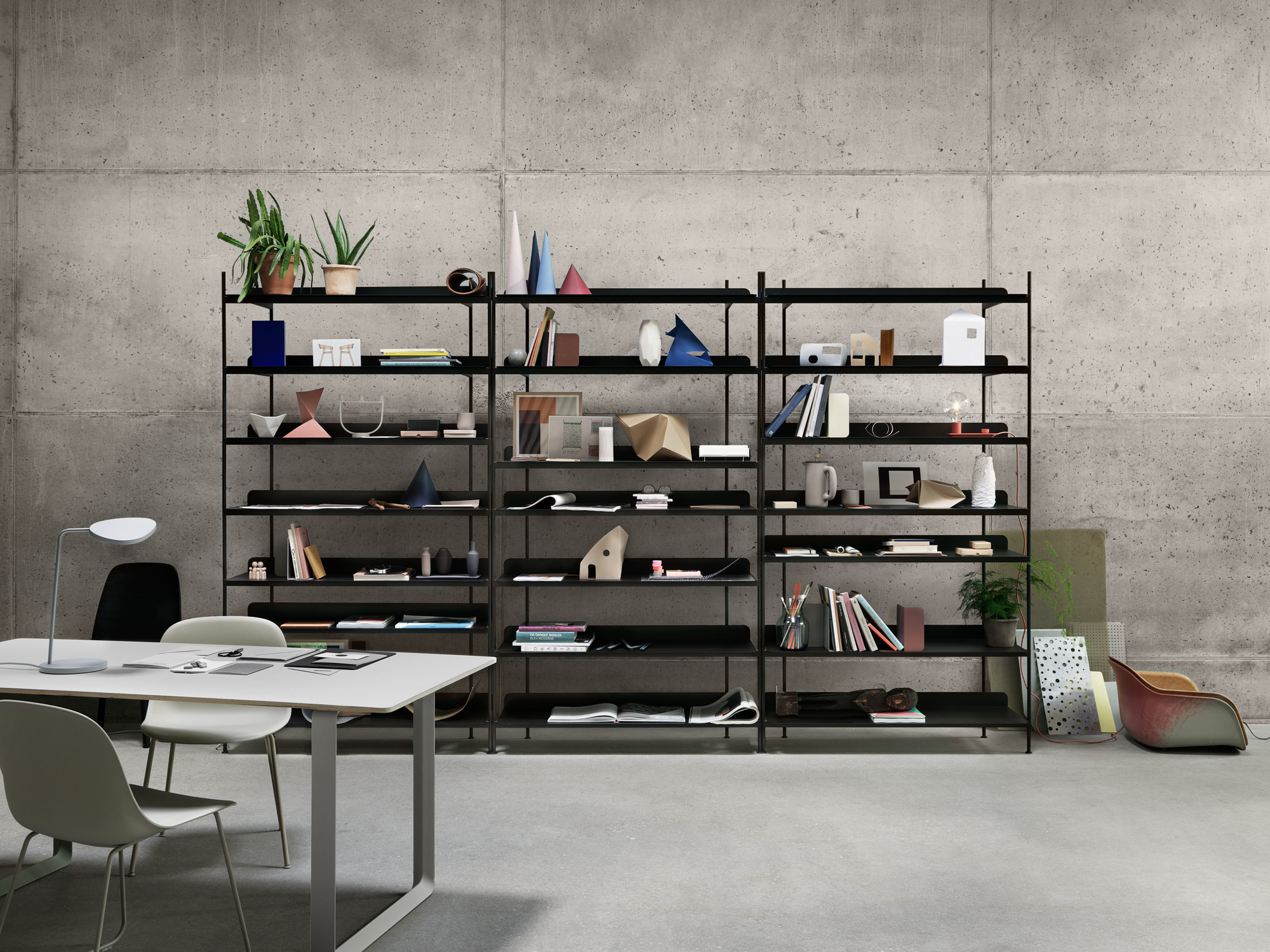 Muuto 70/70 Table Tisch mit Stühlen und Lampe vor einem schwarzen Bücherregal auf Betonwand.