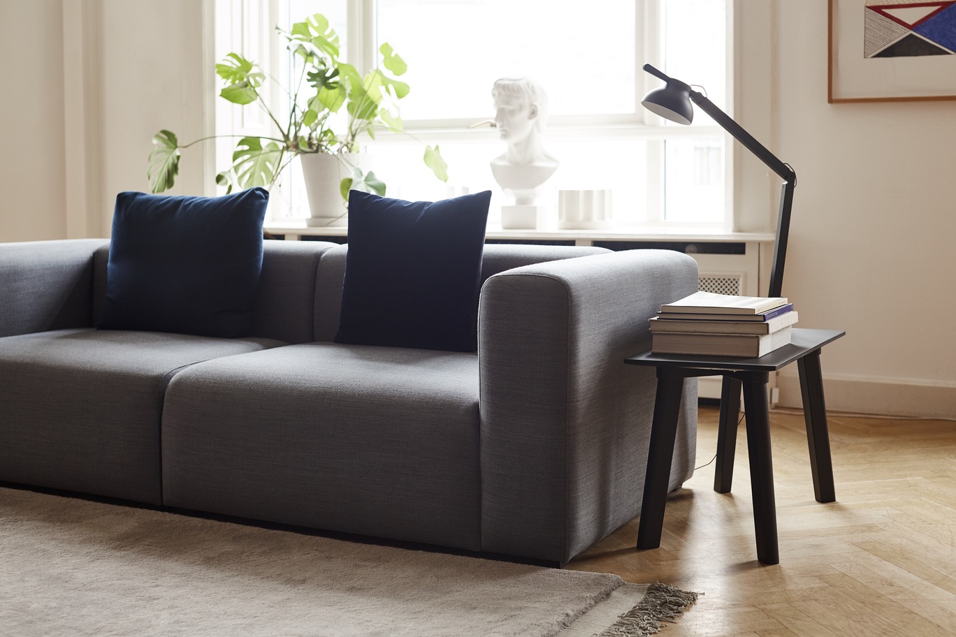 Graues modulares Sofa mit blauen Kissen, Beistelltisch mit Lampe und Büchern in hellem Wohnzimmer.