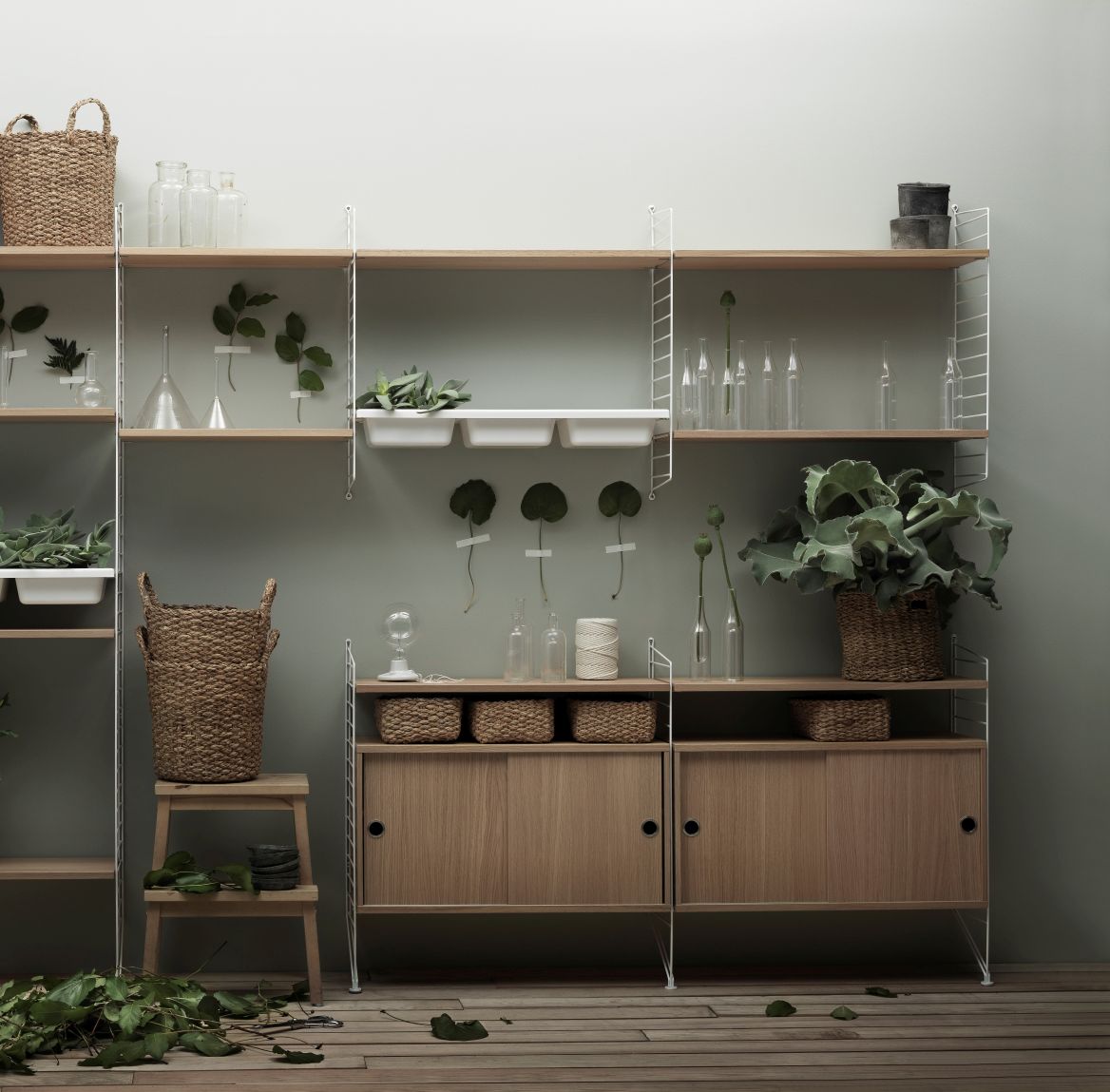 String Furniture Regalsystem mit Walnuss Schrankelement und Schiebetüren, dekoriert mit Pflanzen und Körben.
