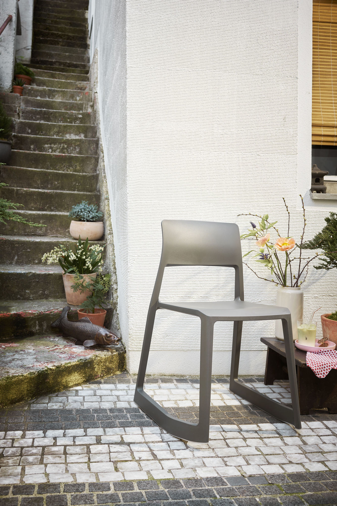 Vitra Tip Ton RE Stuhl in Soft Grey vor weißer Wand und Treppe mit Pflanzen.