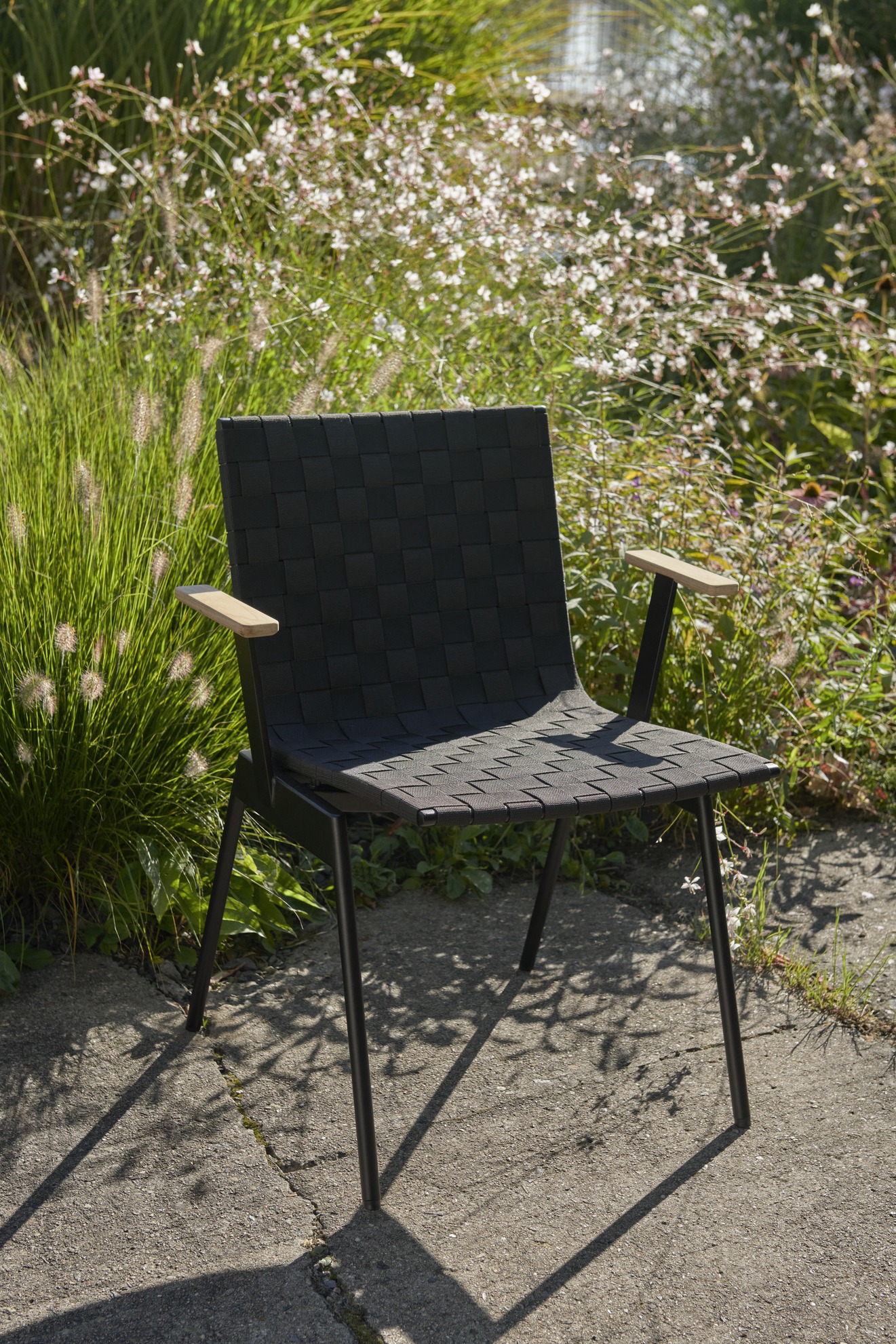 AndTradition Ville AV34 Outdoor Stuhl mit schwarzem, gewebtem Sitz und Armlehnen aus Holz im Garten.