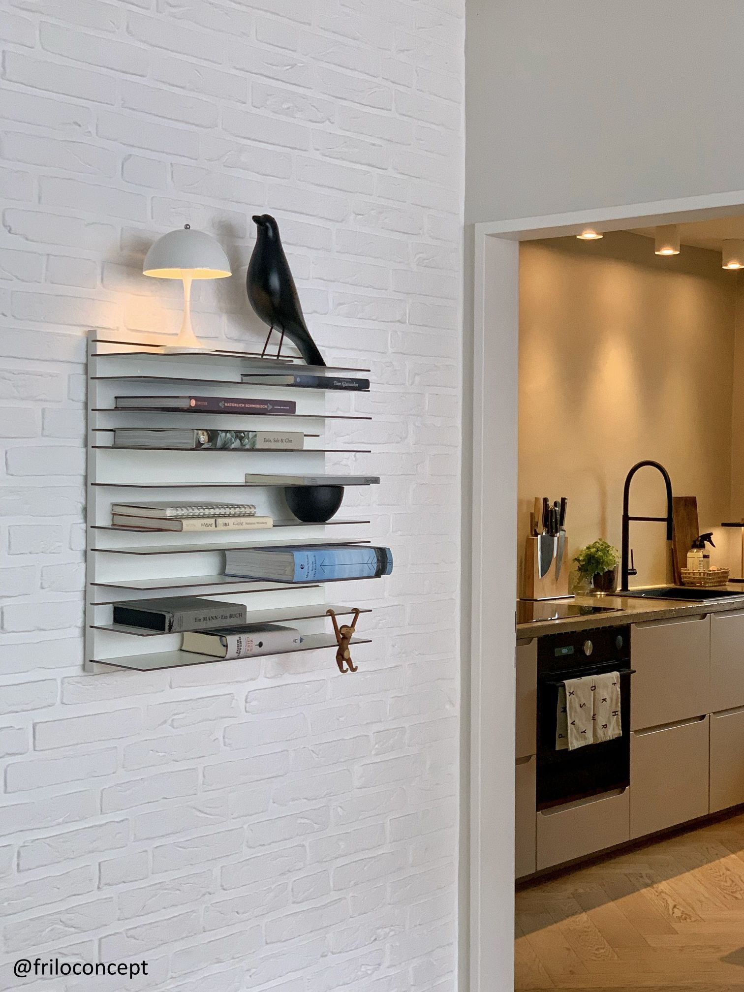 Paperback Bücherregal Spectrum an weißer Ziegelwand, gefüllt mit Büchern, Lampe und Dekoration im modernen Wohnraum.