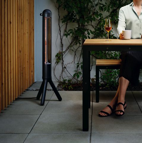 Schwarzer Terrassenheizer mit Infrarot-Technologie neben Frau am Tisch, für gemütliche Abende im Freien.