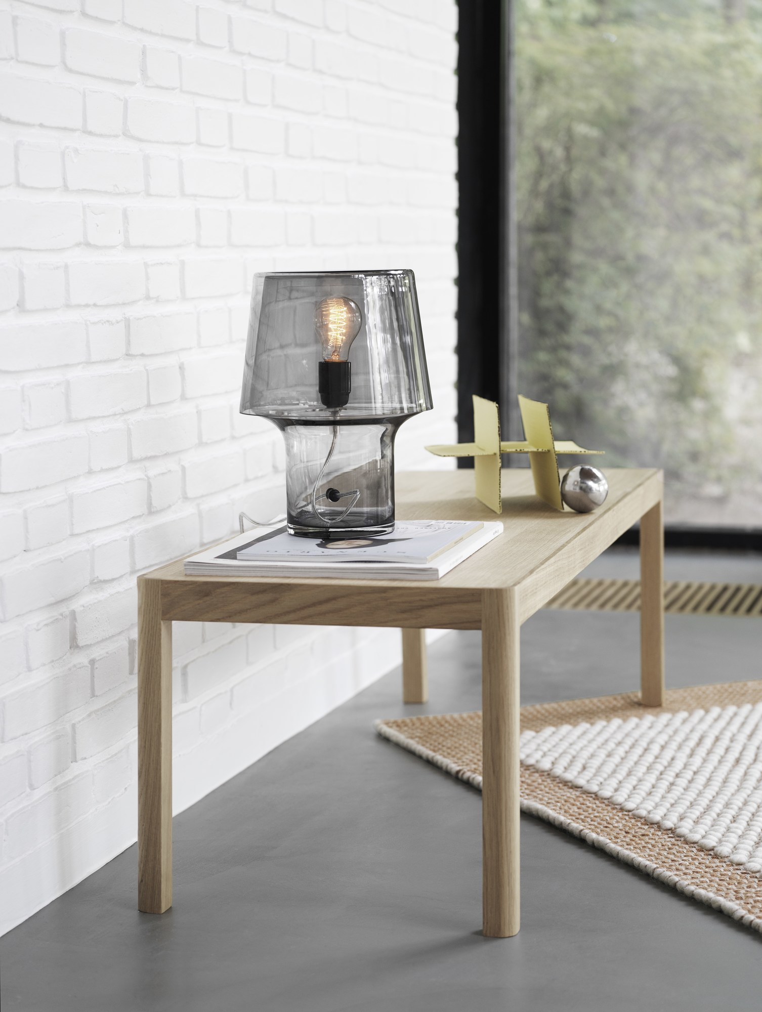 Nahaufnahme des Muuto Pebble Rug Teppichs, teilweise unter einem Holztisch sichtbar, mit grauer Lampe.