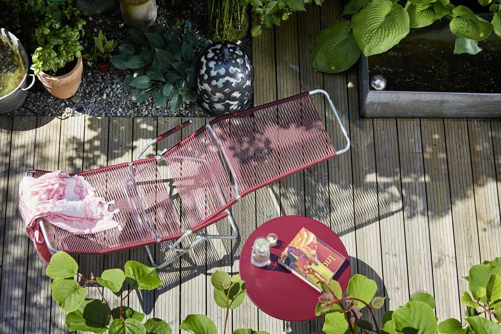 Altorfer Embru Liegestuhl in Verkehrsrot auf Holzterrasse mit Beistelltisch und Gartenbepflanzung. Outdoor Möbel.