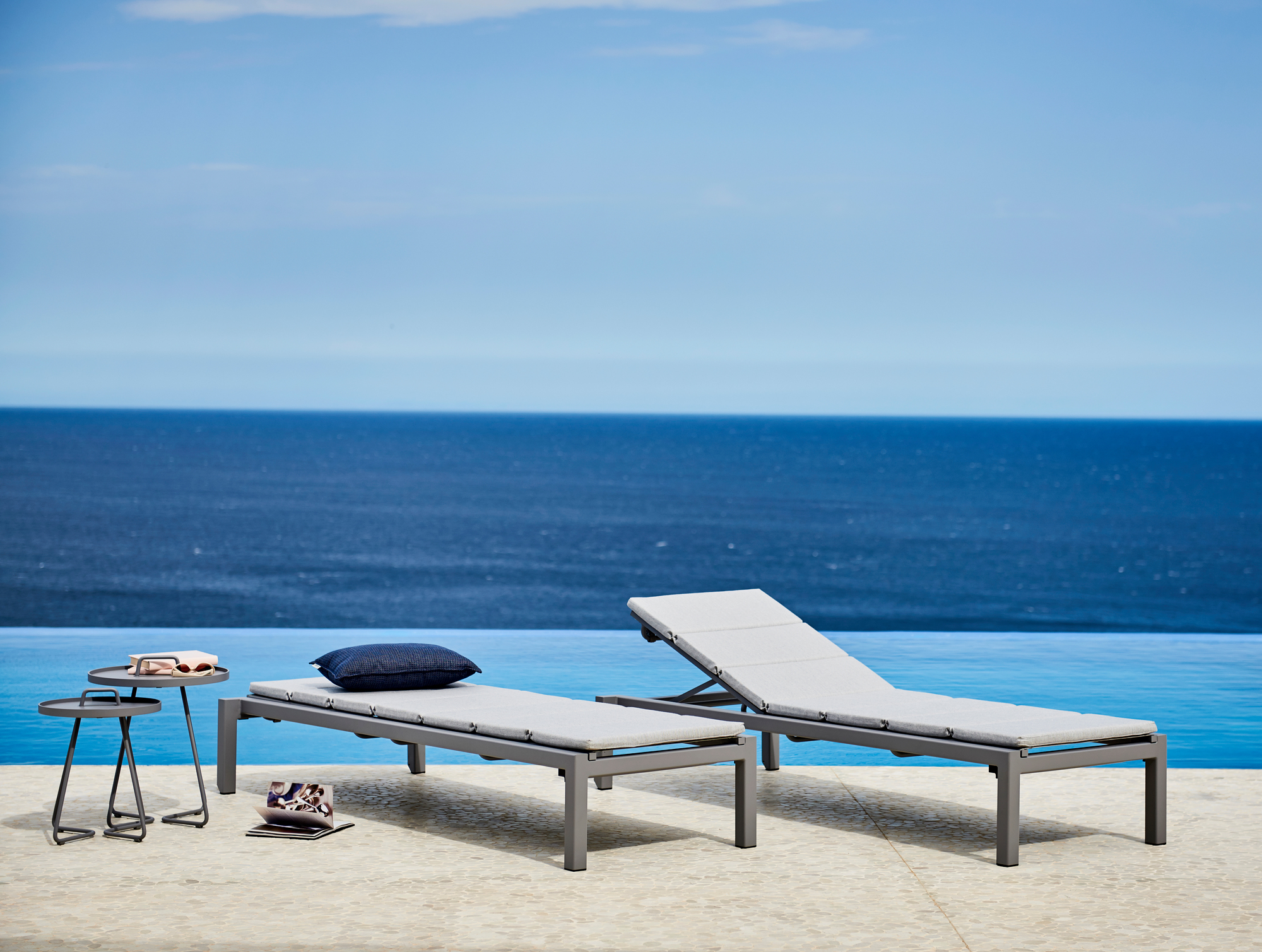 Zwei graue Cane-Line Relax Sonnenliegen mit Kissen und Beistelltisch vor blauem Meer.