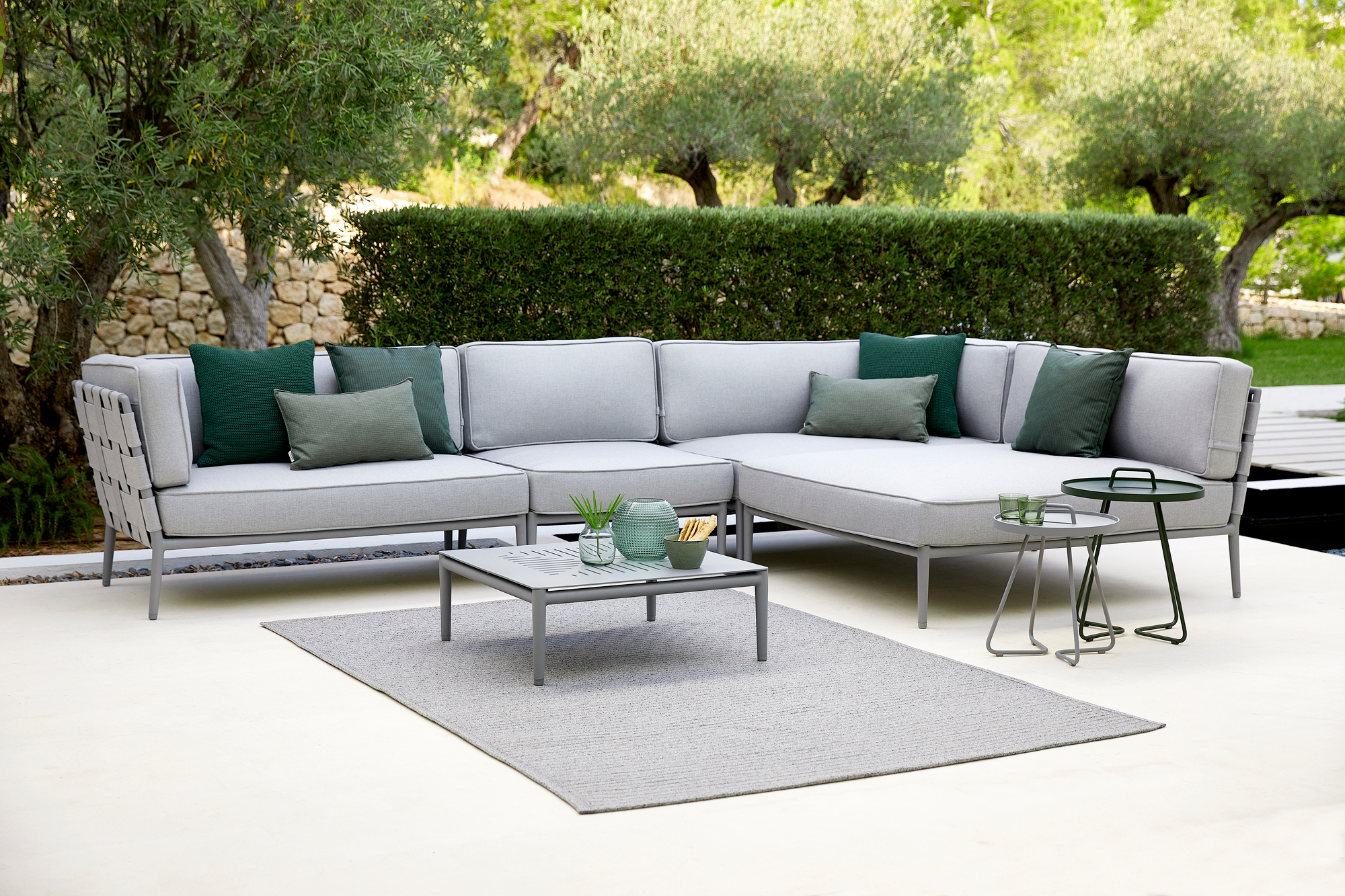 Conic AirTouch Modulsofa von Cane-Line, hellgraues Outdoor Sofa mit grünen Kissen und passendem Gartentisch.