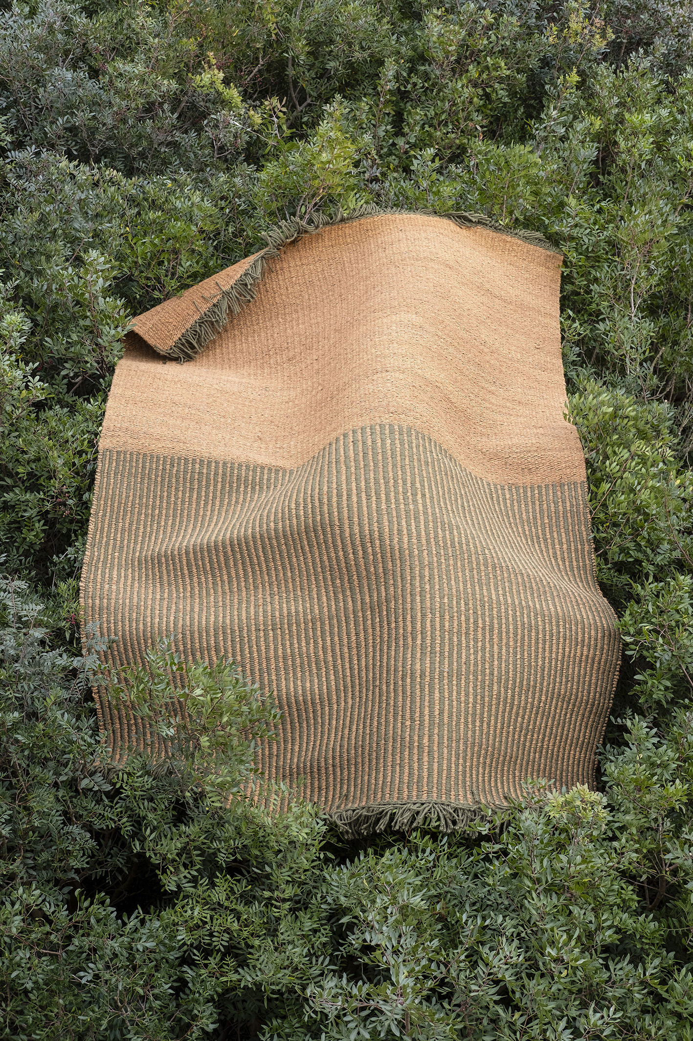 Nanimarquina Garden Outdoor Teppich in Braun und Grün, drapiert über üppigem grünen Buschwerk.