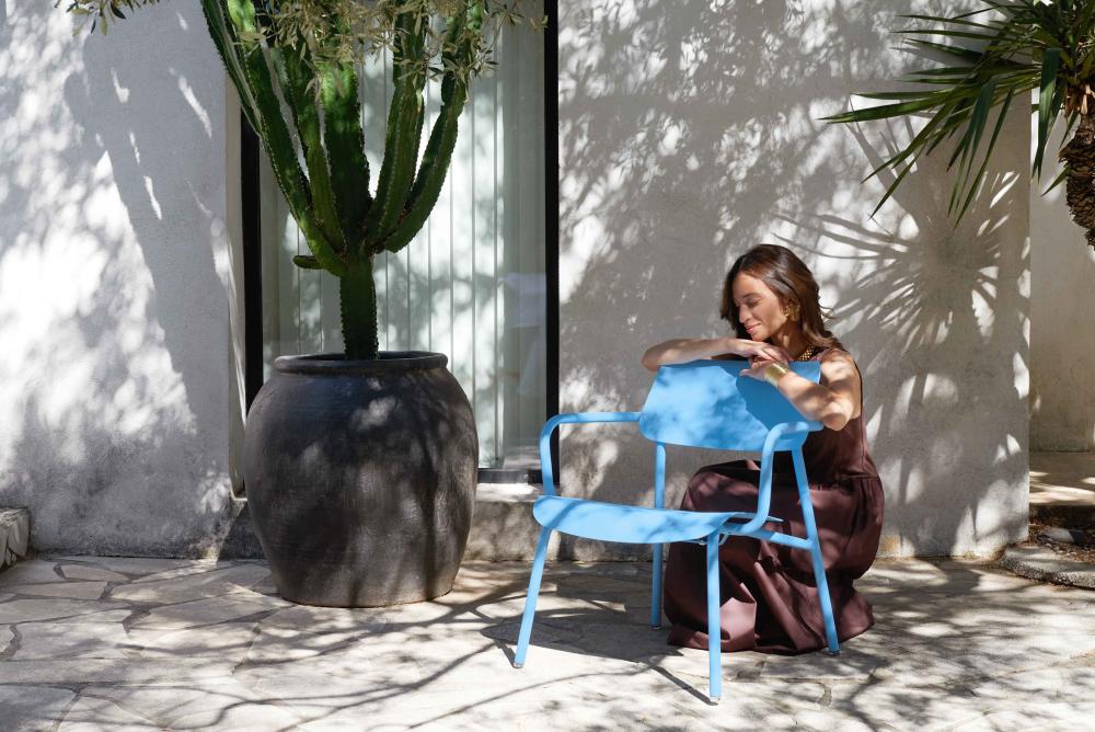 Frau entspannt auf blauem Fermob Studie Lounge Sessel im Freien, umgeben von Schatten und mediterraner Atmosphäre.
