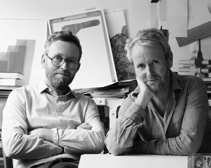 Schwarz-Weiß-Foto von zwei Männern, den Designern des Produkts, im Atelier mit Skizzen im Hintergrund.