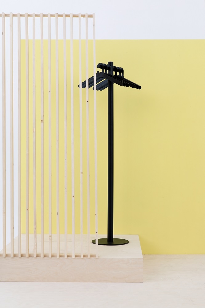 Schwarzer Cascando Pole Garderobenständer mit Kleiderbügeln vor gelbem Hintergrund und Holzpaneel. Moderner Garderobenständer für Büro und Zuhause.