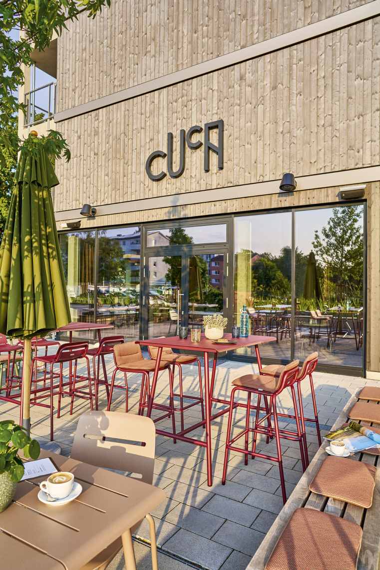 Weishäupl Vapio Hochtisch Outdoor in Rot vor modernem Gebäude mit Außenbereich und Sitzgelegenheiten.