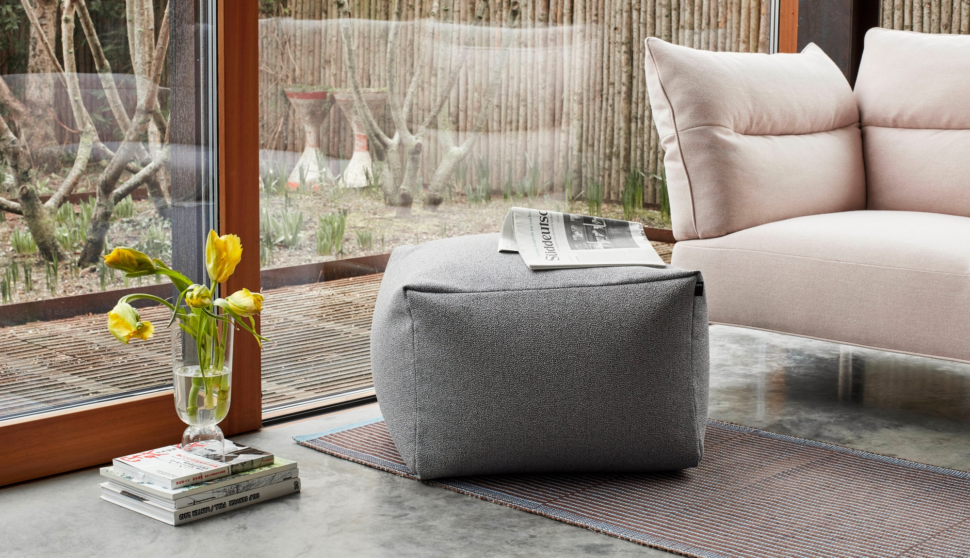 Grauer Hay Teppich, ein gestreifter Teppich, dient als Unterlage für einen grauen Sitzhocker im Wohnzimmer.
