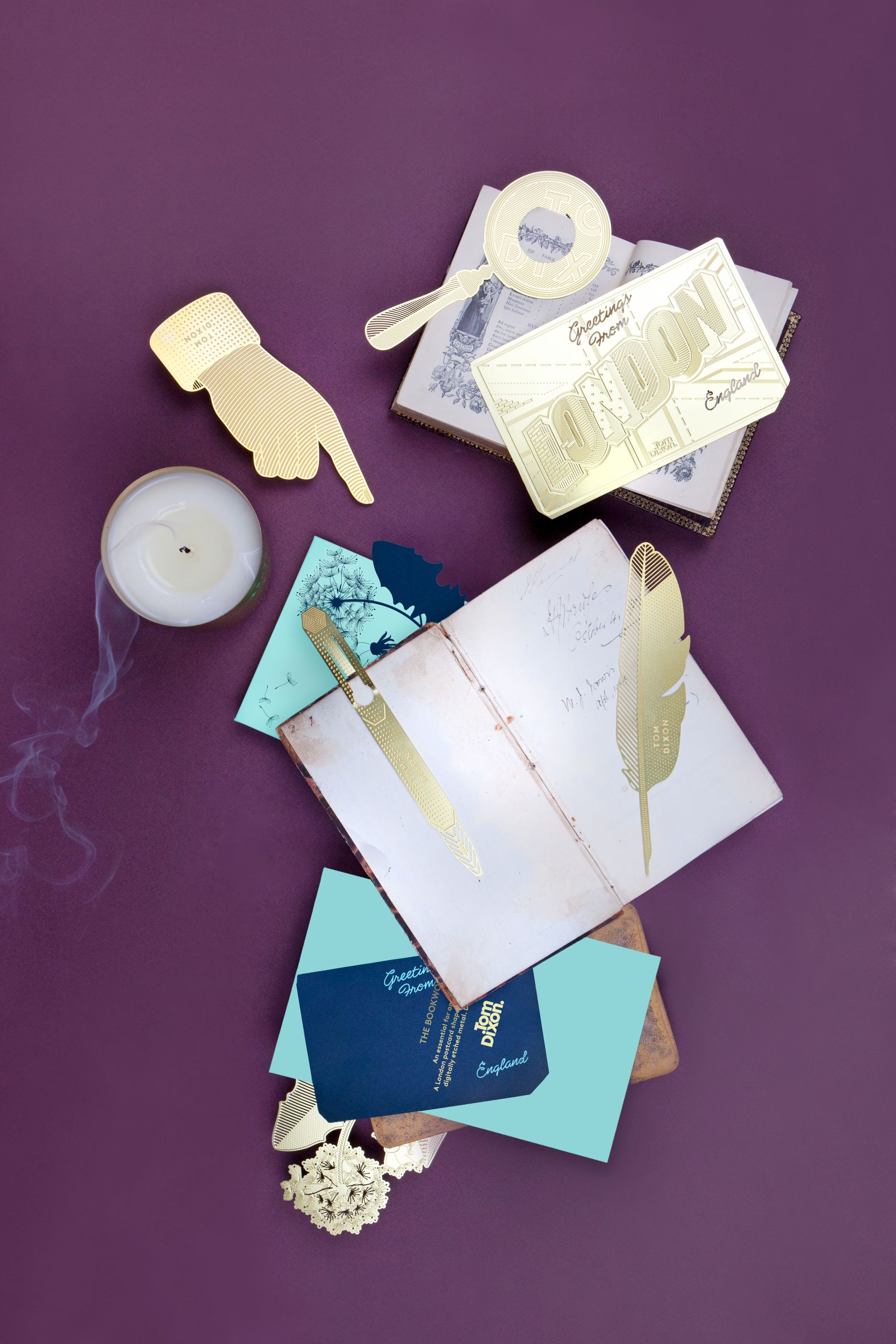 Produktfoto: Tom Dixon Briefbeschwerer, Lesezeichen und Postkarten aus goldfarbenem Metall auf lila Hintergrund.