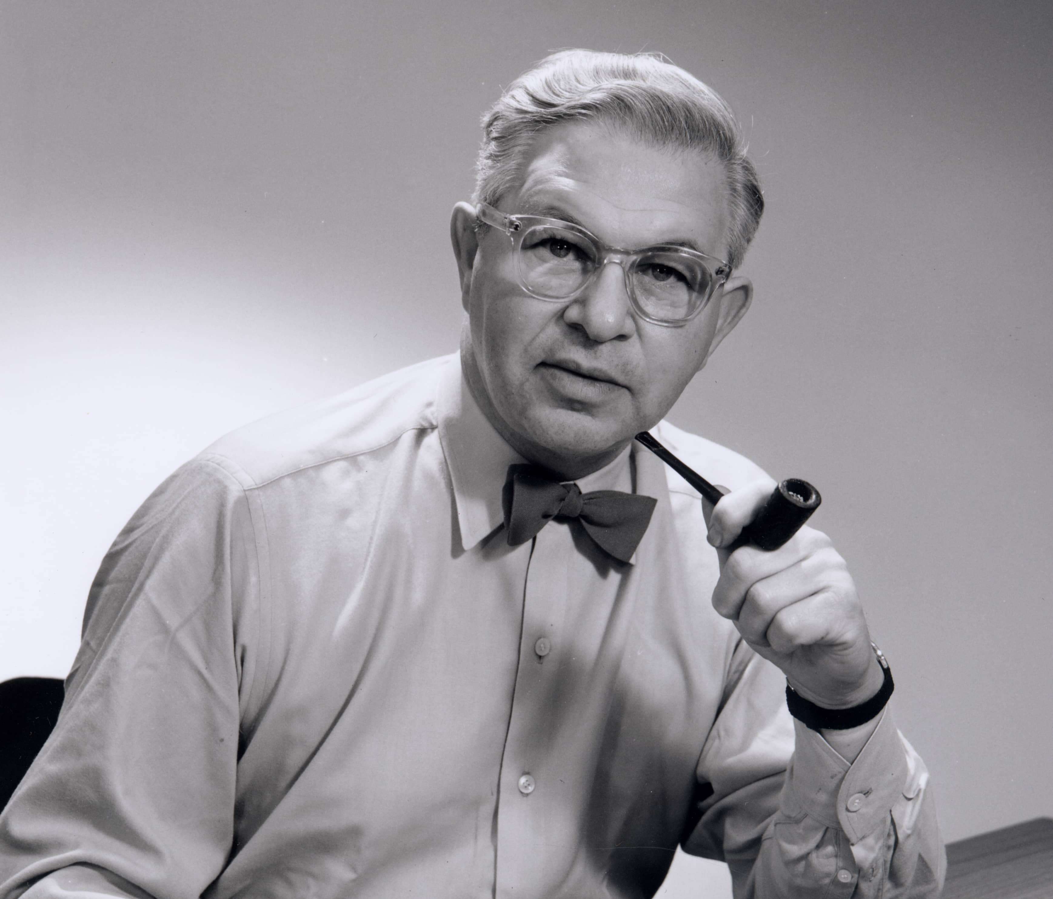 Schwarz-Weiß-Foto eines Mannes mit Brille, Fliege und Pfeife. Klassischer Stil.
