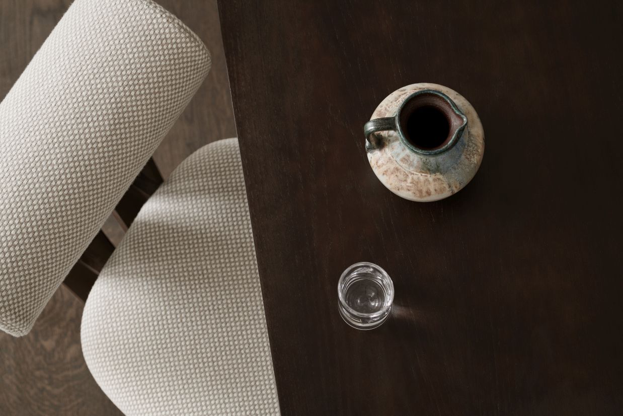 Detailaufnahme des NORR11 Hippo Stuhls mit beigem Stoffbezug, dunklem Holztisch, Krug und Glas.