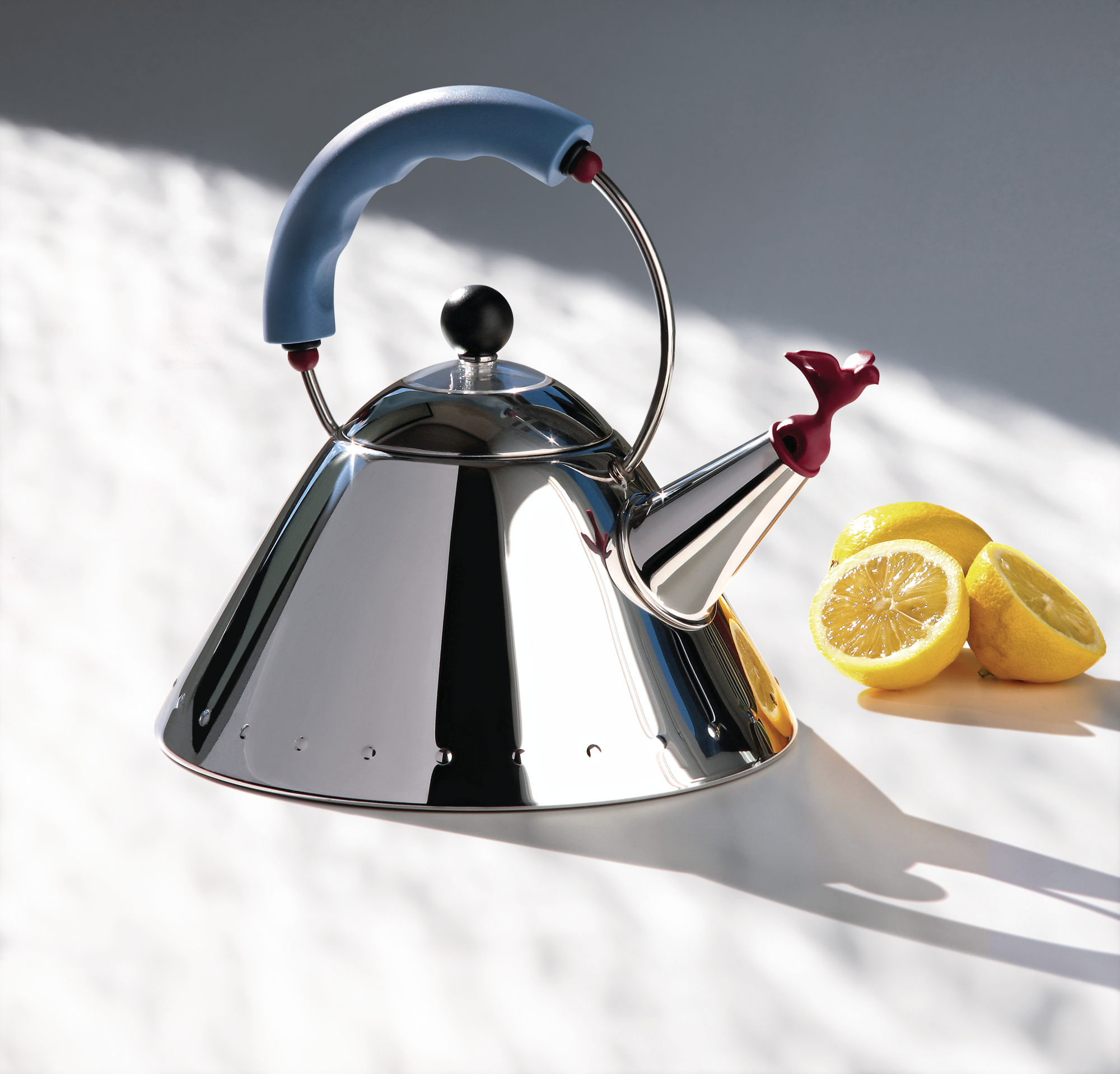 Alessi Wasserkessel 9093 aus Edelstahl mit blauem Griff und Vogelpfeife neben Zitronen.