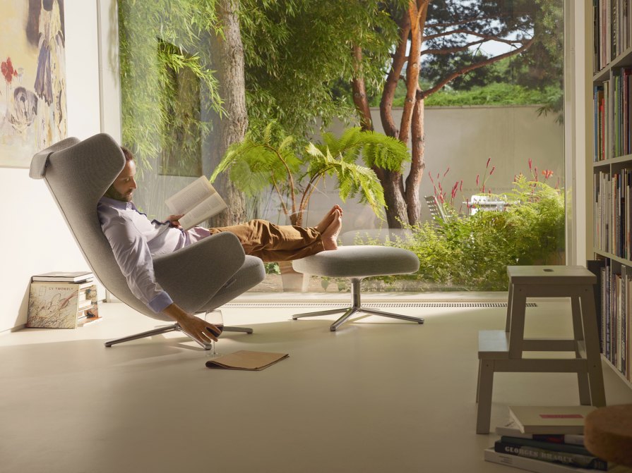 Mann entspannt im grauen Vitra Grand Repos Sessel mit Ottoman, liest Buch, Glas Wein.