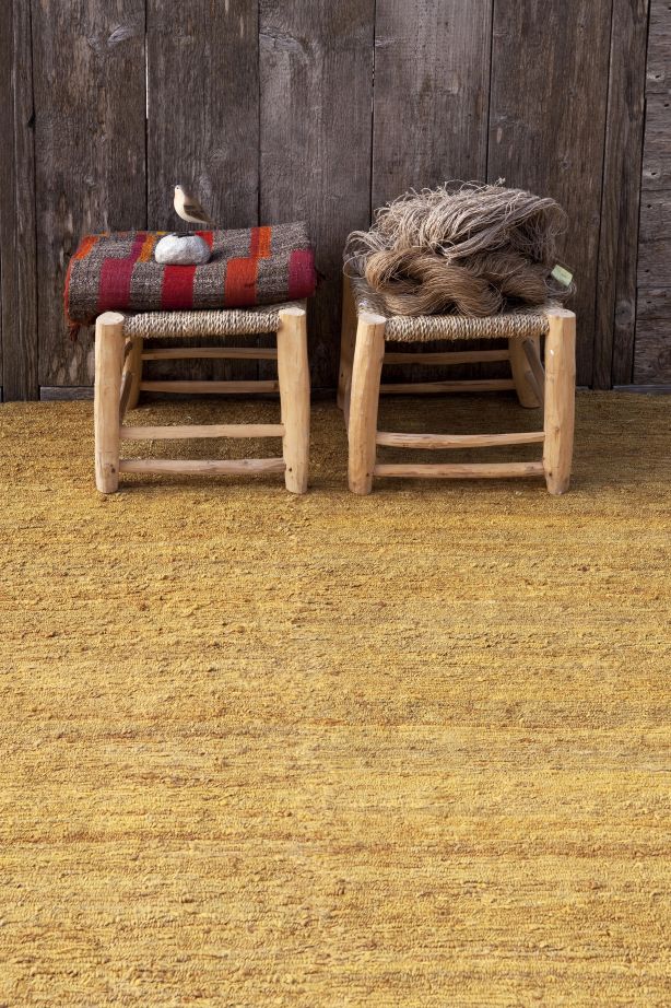 Nanimarquina Earth Rug Teppich, goldener Wollteppich mit zwei Hockern vor einer Holzwand.