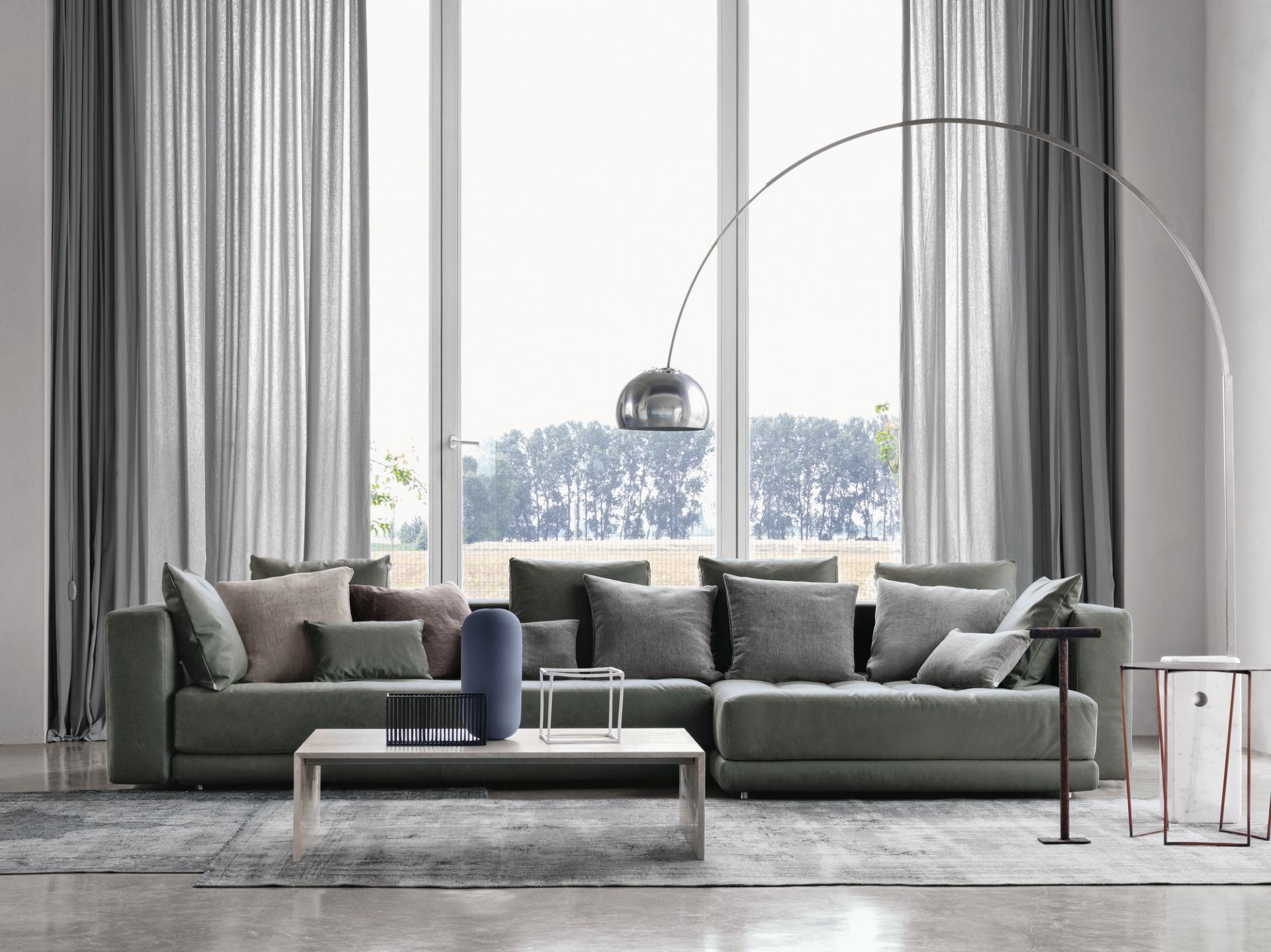 Großes, graues Ecksofa mit vielen Kissen, Couchtisch und Stehlampe vor einem Fenster mit Vorhängen.