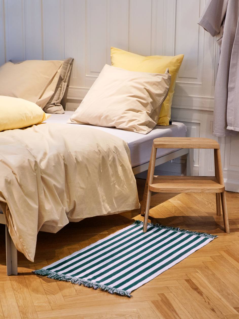 Heller Raum mit Bett, Teppich und dem Hay Butler Hocker aus hellem Holz.