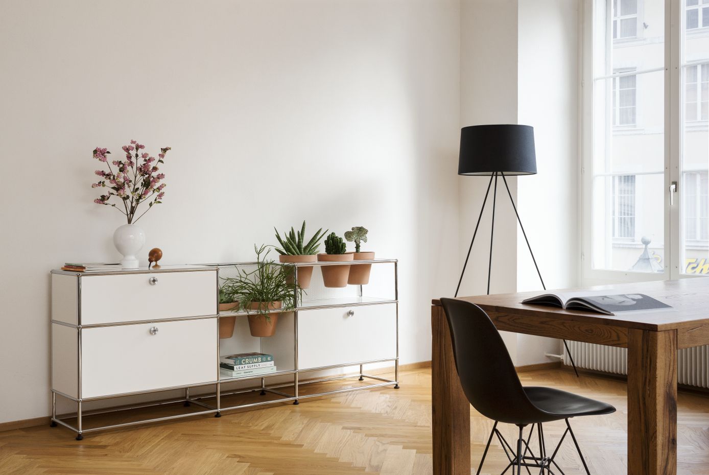 Modernes weißes Sideboard mit Chromgestell, Pflanzen, Tisch mit Lampe und Stuhl in hellem Raum.