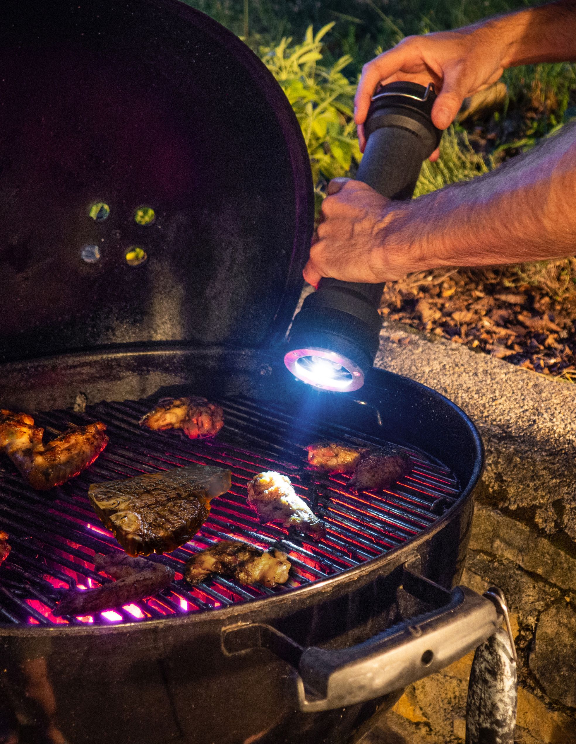 Mann grillt Steaks und Hähnchen auf einem Holzkohlegrill und leuchtet mit einer Taschenlampe.
