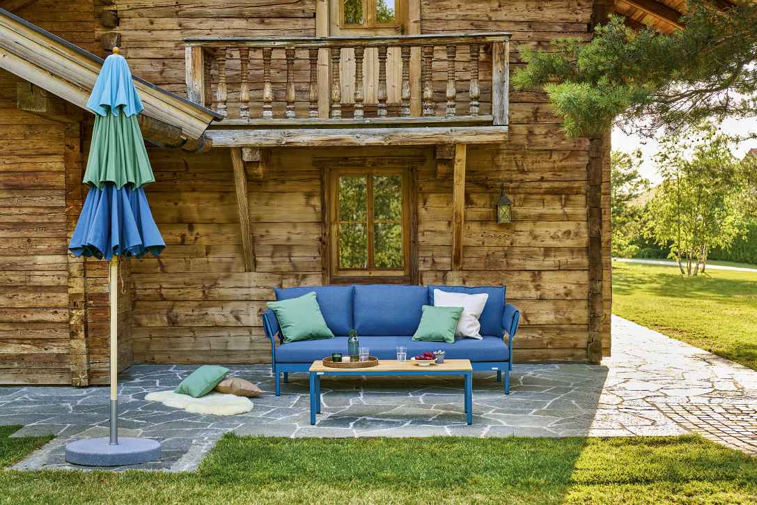 Weishäupl Klassiker Schirm in Blau-Grün neben blauer Outdoor-Couch vor rustikalem Holzhaus.