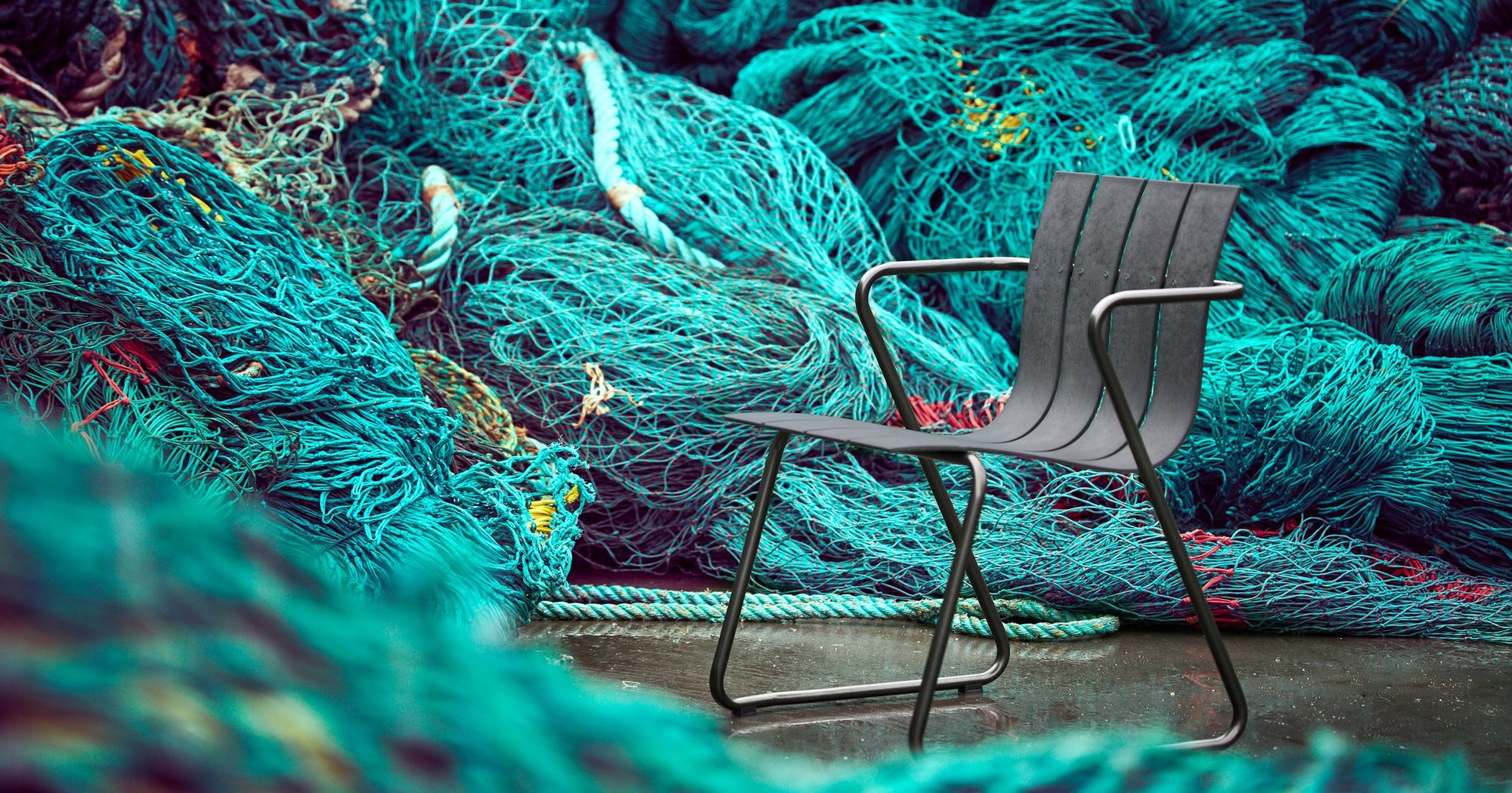 Ocean Chair Outdoor Stuhl in Sand vor blauem Fischernetz. Nachhaltiger Gartenstuhl für den Außenbereich.