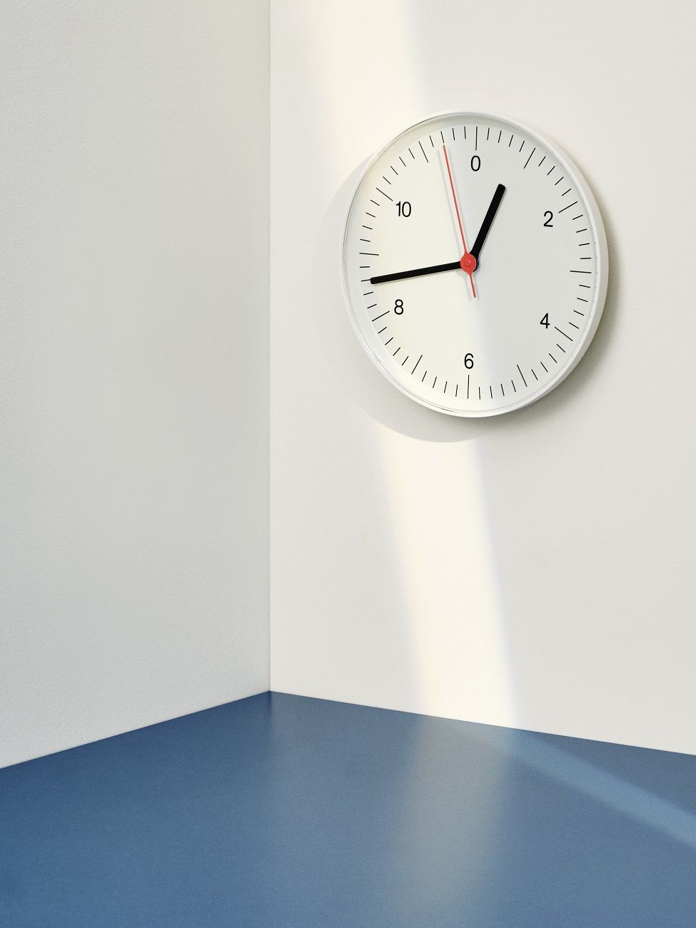 Weiße Hay Wanduhr mit minimalistischem Design an einer weißen Wand über einem blauen Boden.