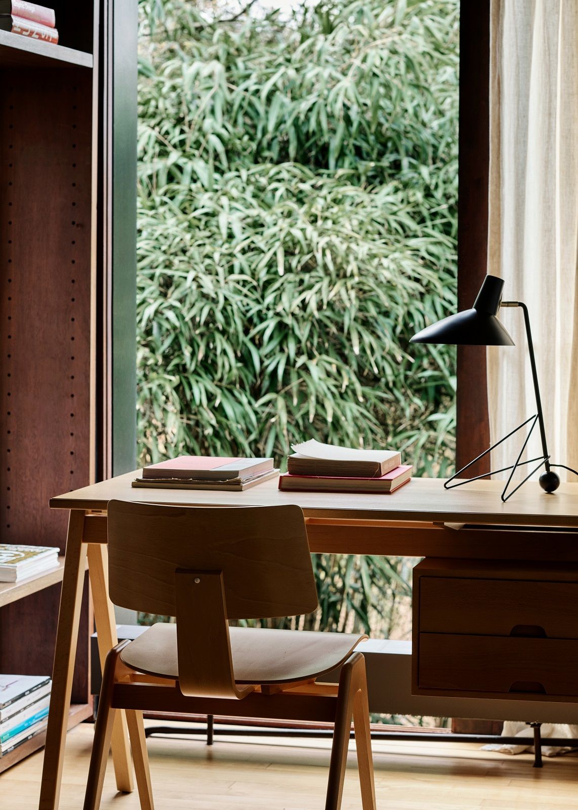 AndTradition Daystak Tisch mit Holzstuhl und schwarzer Lampe vor Fenster mit Grünpflanzen.