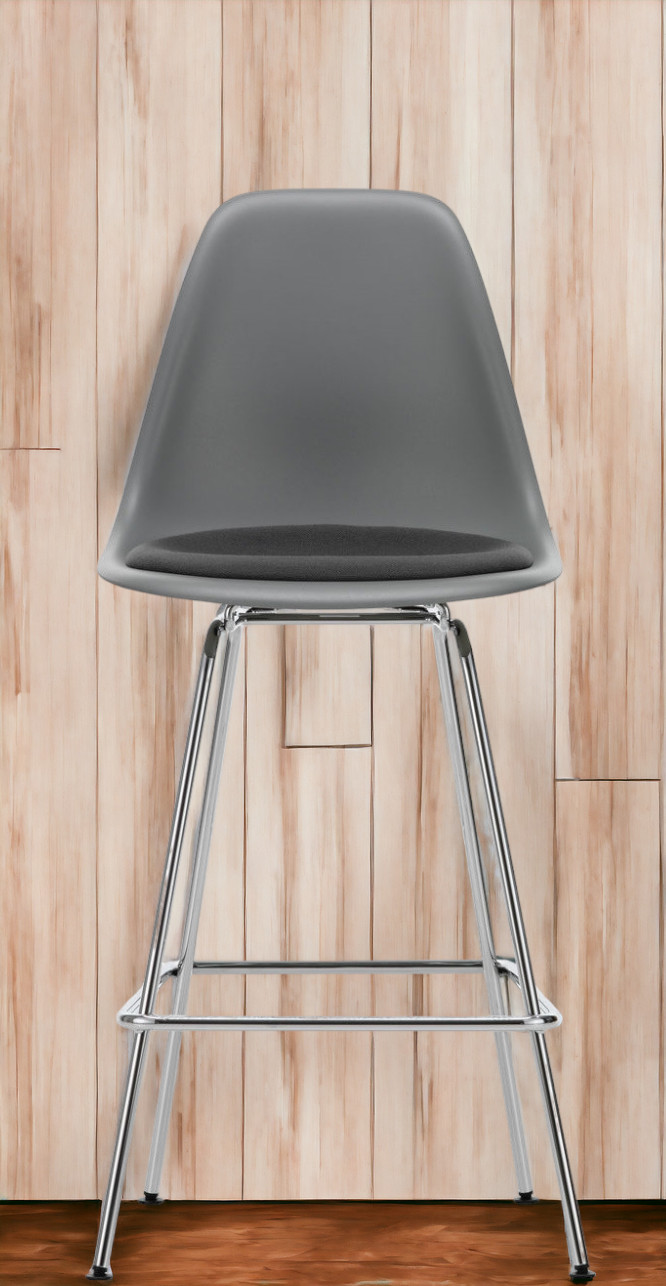 Grauer Eames Plastic Bar Stool Barhocker von Vitra mit Sitzpolster und verchromtem Gestell vor Holzwand.