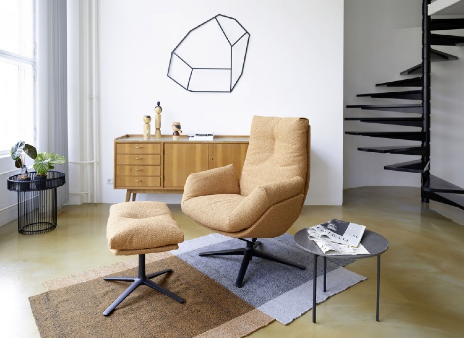Beiger Sessel mit Hocker und schwarzem Drehfuß im modernen Wohnzimmer mit Treppe und Kommode.