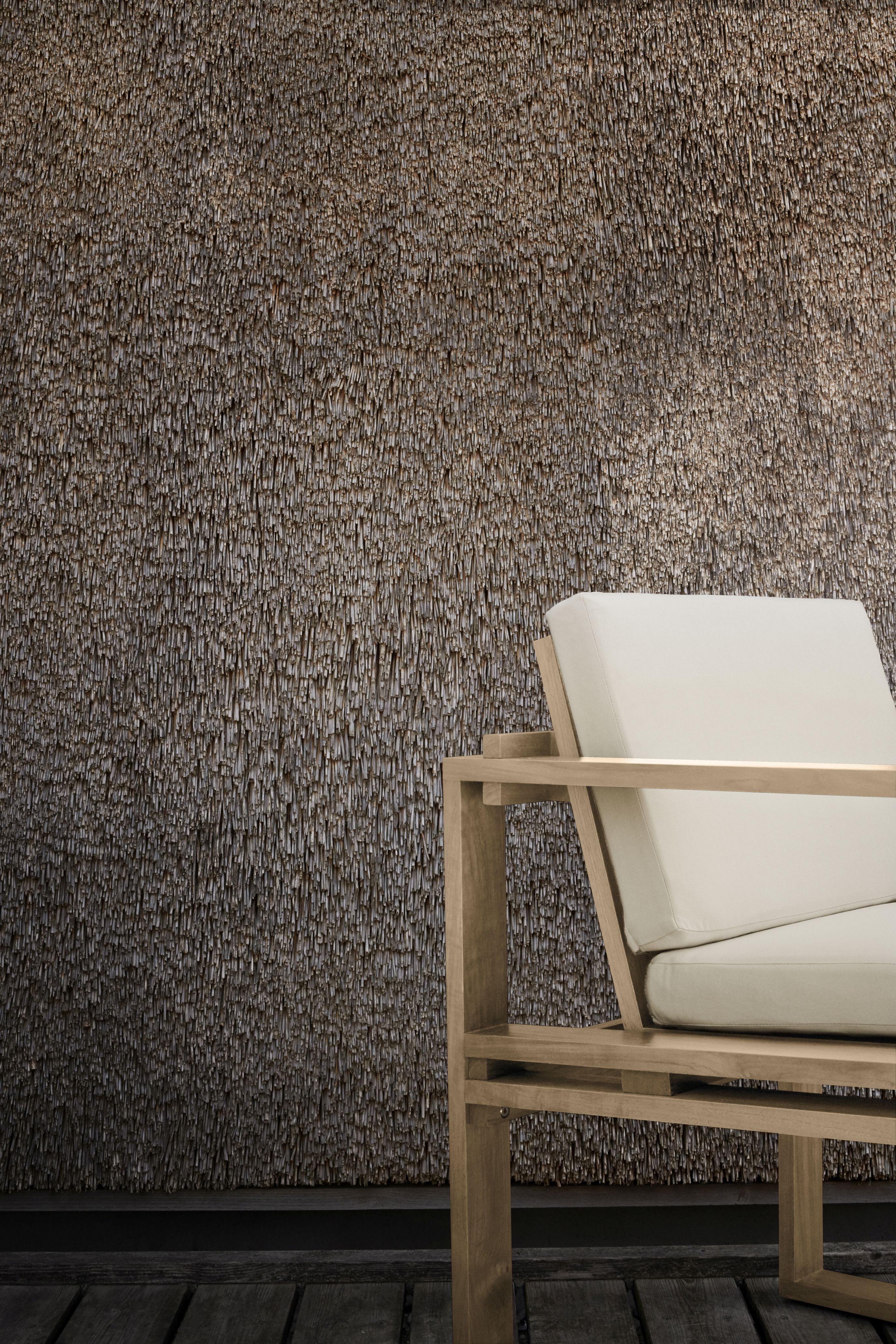 Bodil Kjaer Outdoorstuhl von Carl Hansen, Teakholz mit cremefarbenen Polstern vor einer Reetwand.