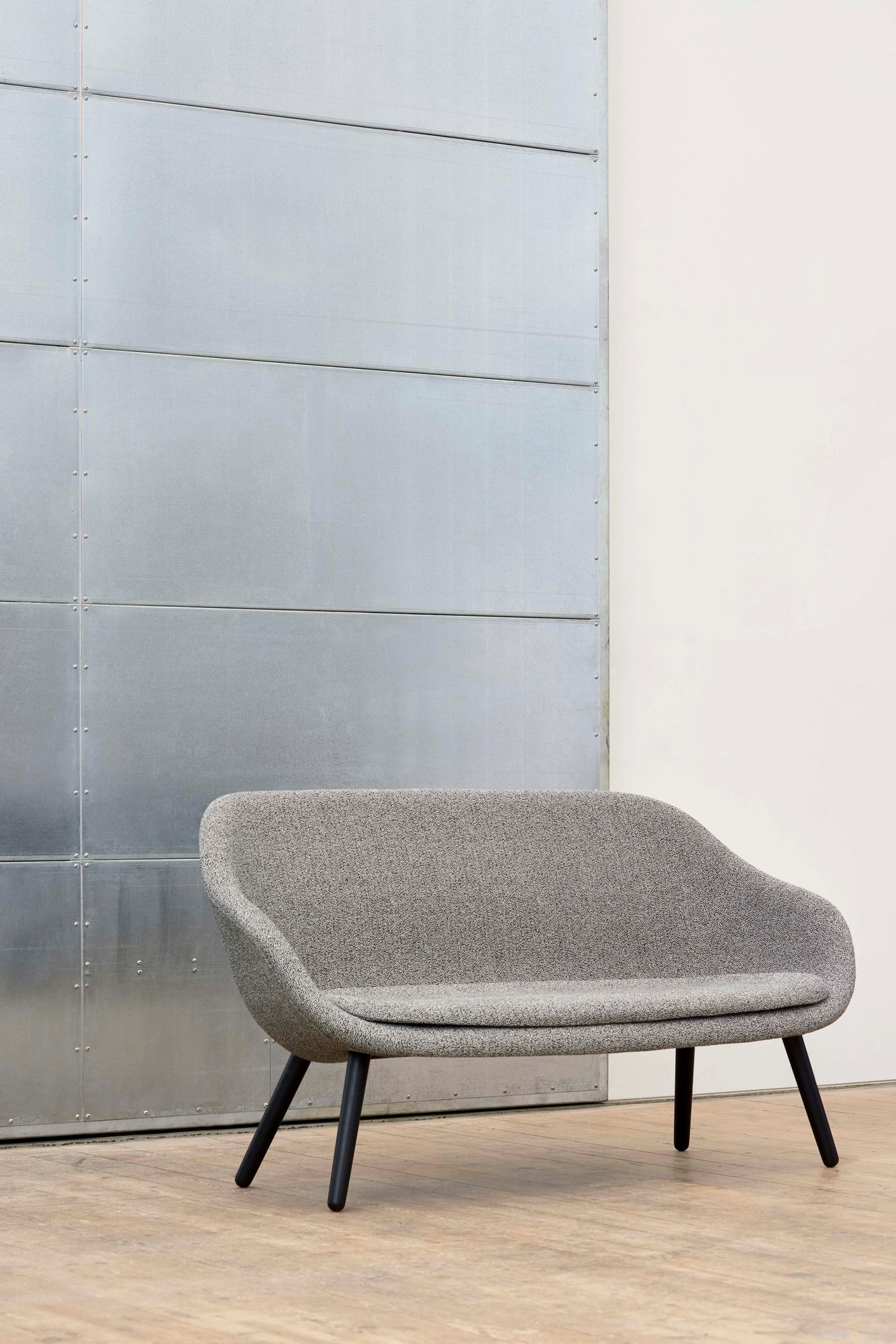 Graues Hay About a Lounge Sofa AAL mit schwarzen Holzbeinen vor Metallwand.