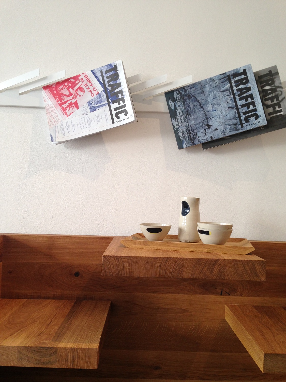 Modernes Interieur mit Holzbank, Serviertablett mit Keramikschalen und Magazinen an einer weißen Wand.