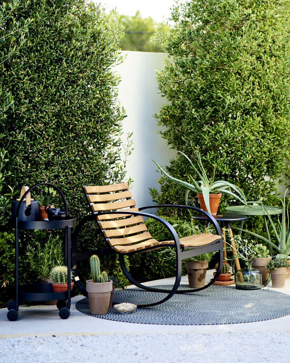 Cane-Line Parc Schaukelstuhl aus Teakholz und schwarzem Metall im Garten mit Pflanzen und Beistelltisch.