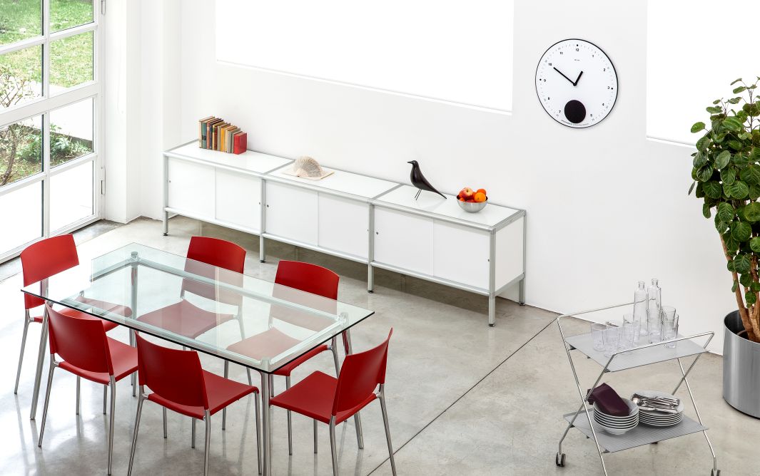 Helle Esszene mit Rexite Appuntamento Wanduhr, Glas-Esstisch, roten Stühlen und weißem Sideboard für modernes Wohnen.
