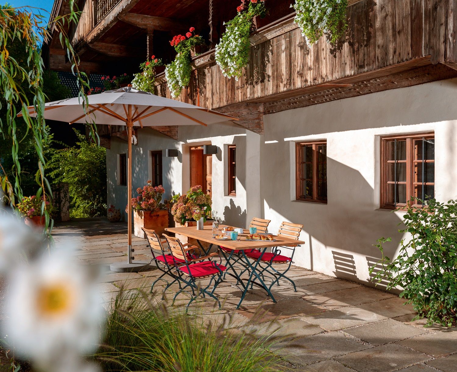 Weishäupl Teakschirm spendet Schatten über einer Gartengarnitur mit Holztisch und roten Stühlen auf einer Terrasse.