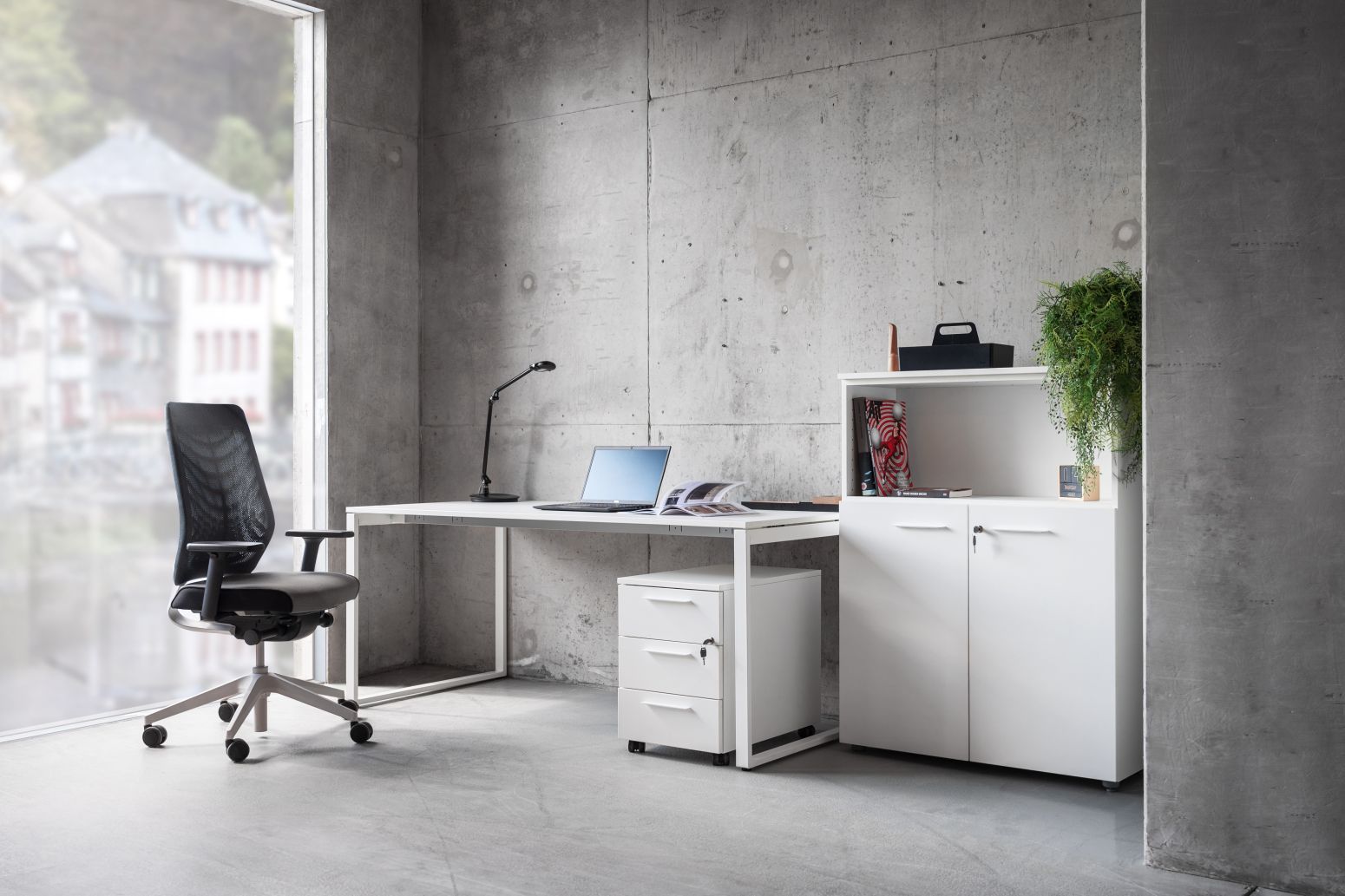 Modernes Büro mit weißem Schreibtisch, Rollcontainer, Büroschrank und ergonomischem Bürostuhl vor Betonwand.